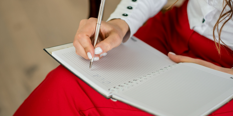 A woman in bright red pants taking notes with a pen in a notebook